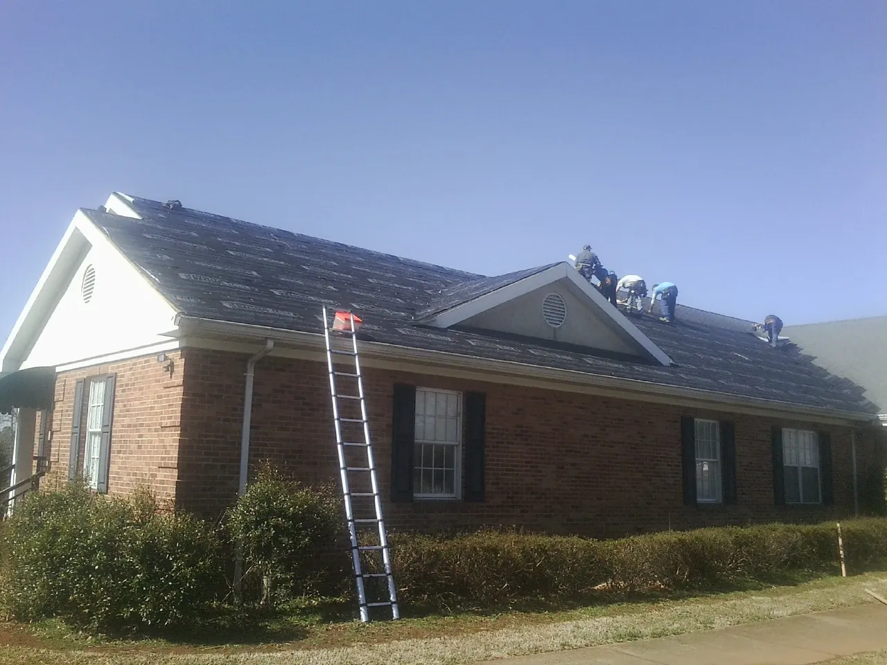 Crew tearing shingles off a brick ranch roof with a ladder set — Metal Roof Repair work by our crew in Rockingham