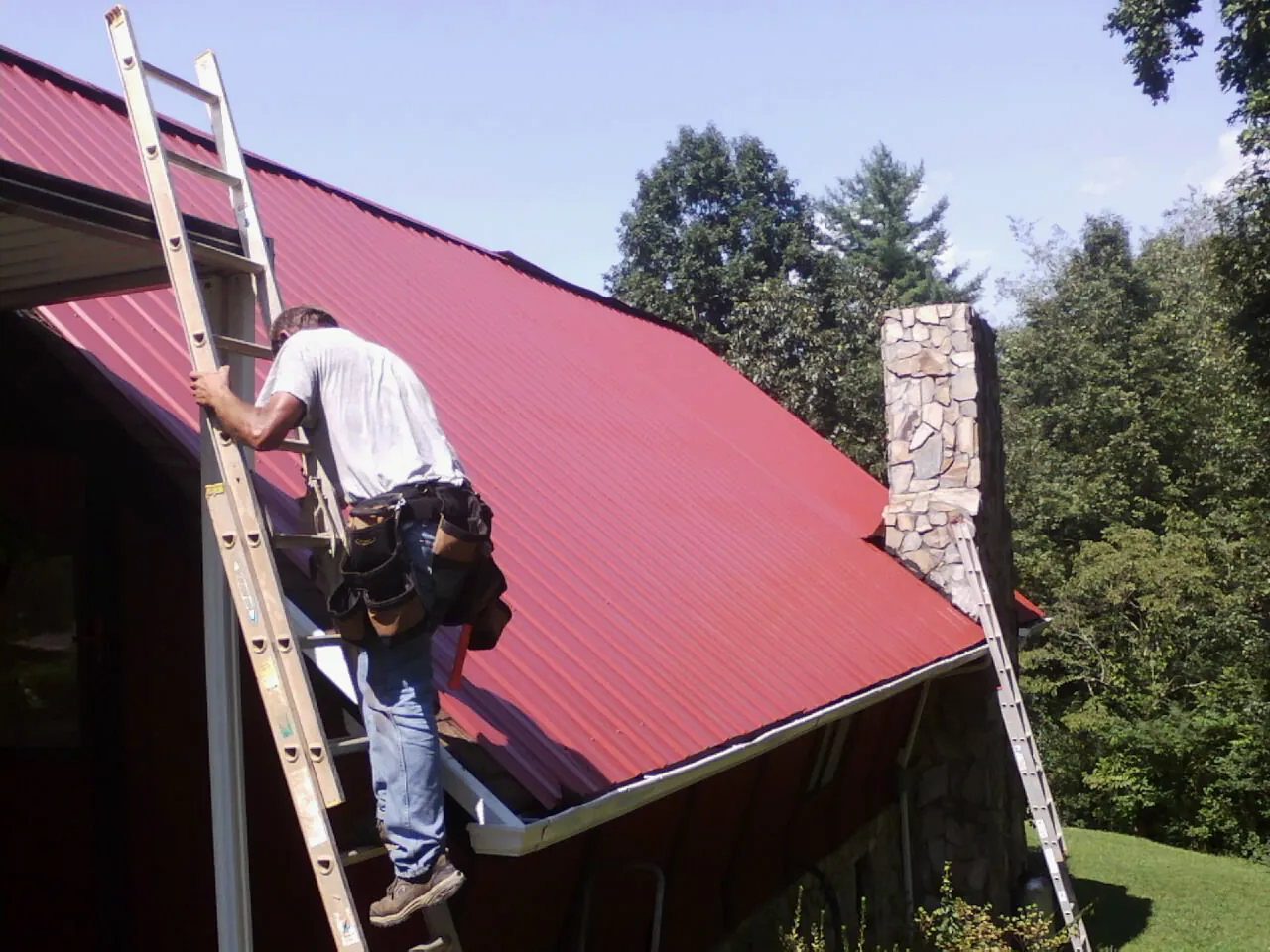 Roofer on a ladder beside a red metal roof and stone chimney — Soffit & Fascia Repair by Roofing Brothers in Rockingham